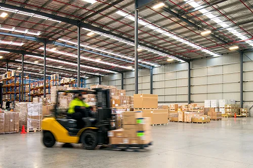 A yellow forklift operator moving goods through a large, high-ceiling industrial warehouse and logistics facility.